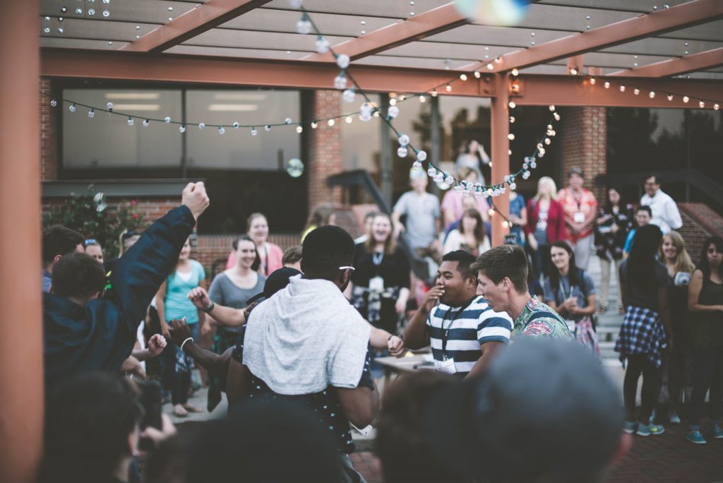 A photograph of an outdoor gathering of about 20 people. Attendees are clustered around tables, some are raising glasses in a toast, and fairy lights hang from the terrace roof.