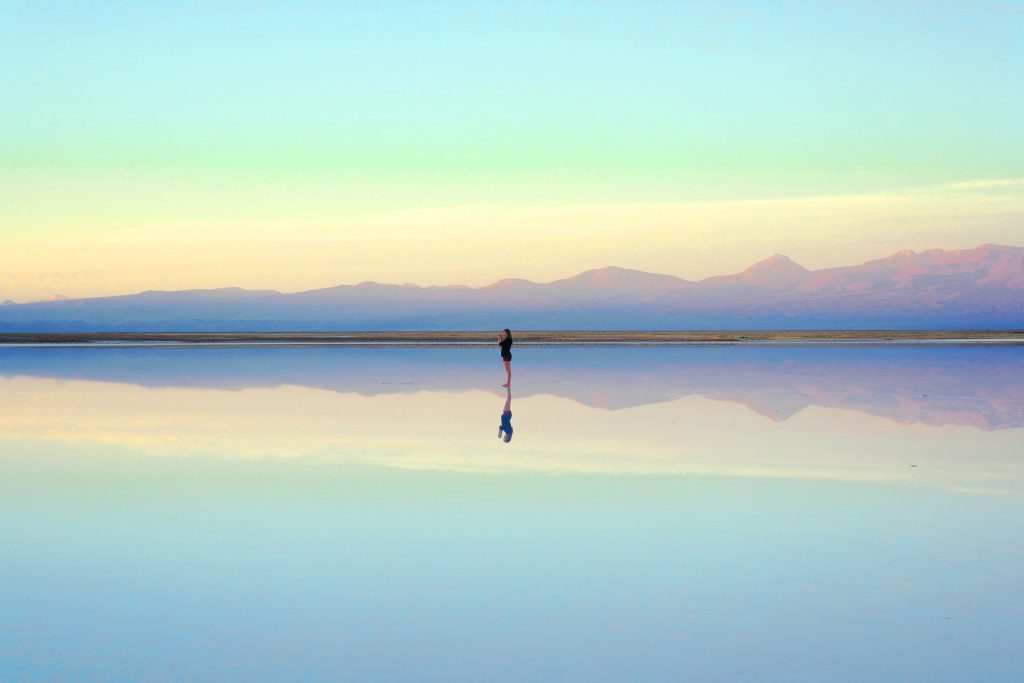 A photo of an individual standing on a wet beach at twilight. The sky is reflected in the surface under their feet. 