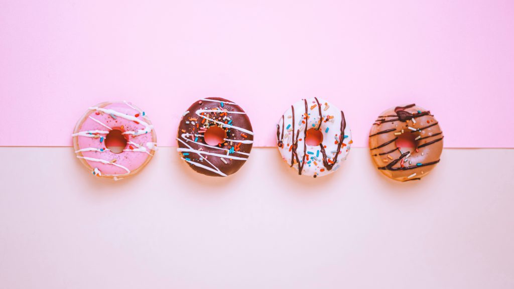 Four frosted ring donuts in a horizontal line, on a surface covered in two pieces of pink paper. 