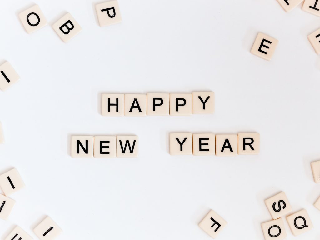 The words "Happy New Year" spelled out in Scrabble tiles. The tiles are off white, the background is white, and there are a few other tiles scattered around the edges of the image. 