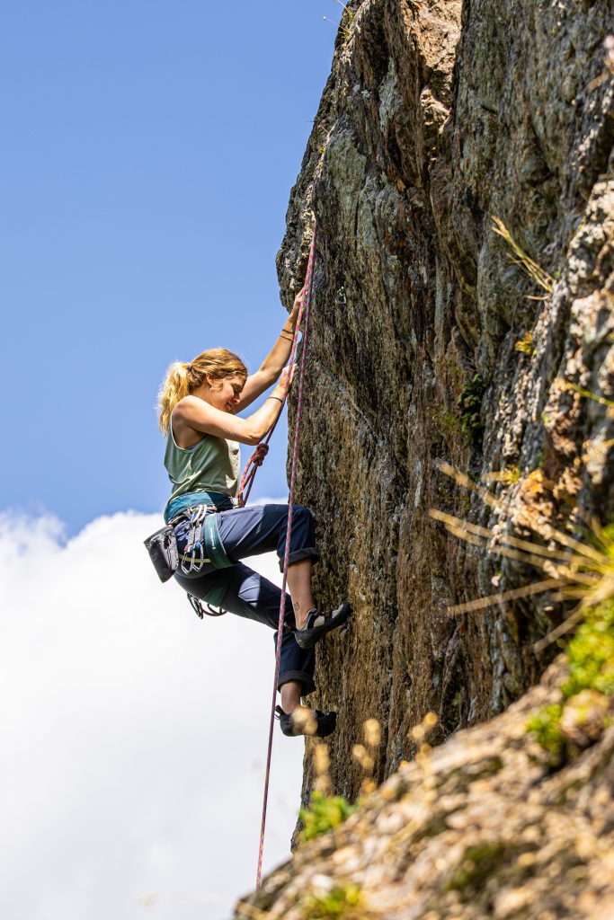 A blonde woman wearing a light green top and blue pants rock climbing outside. 