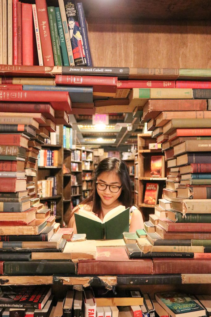 An asian woman wearing glasses reads a green book, surrounded by more books that are arranged in a circle to frame her face. 