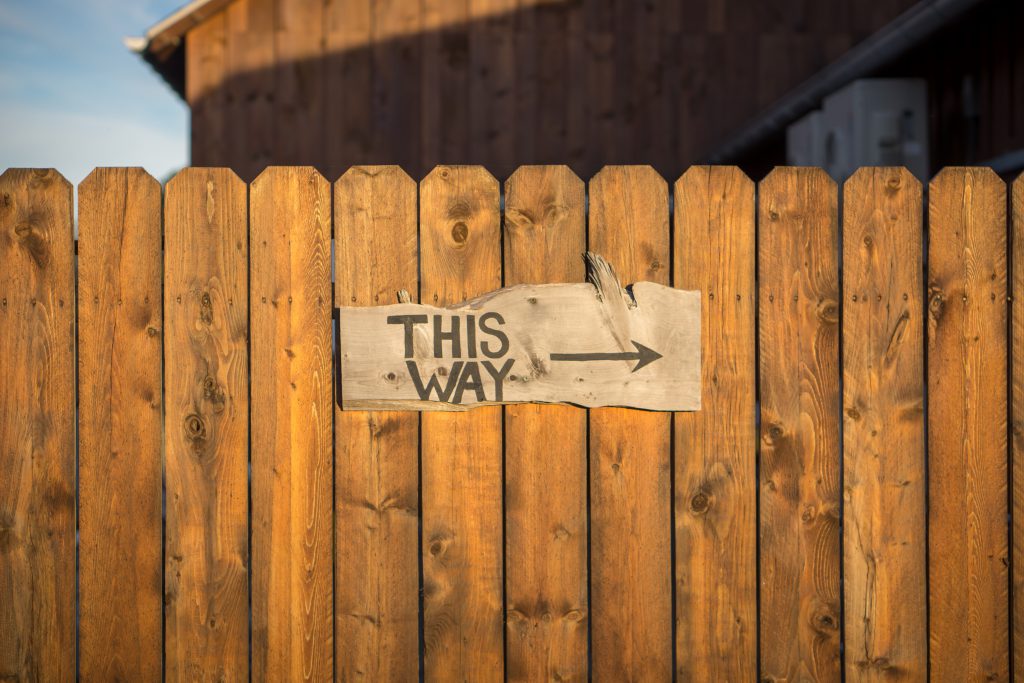 A cardboard sign pinned to a brown wood fences reads "this way," with an arrow pointing to the right. 