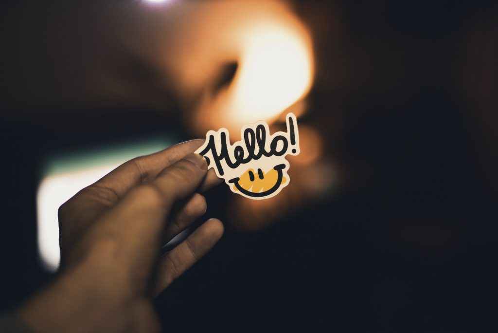 A photo of a hand holding a sticker that says "Hello!" The sticker includes black text on a white background, with a sketch of a smiley face on a scribble of yellow. 