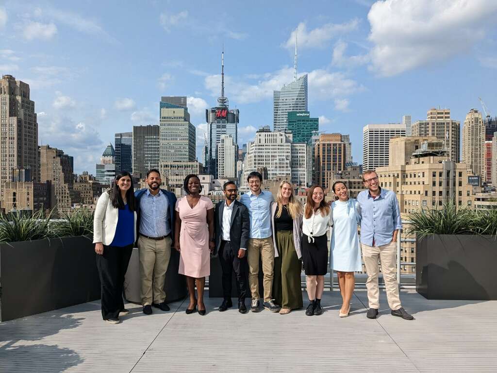 9 individuals stand in a row, smiling with their arms around each other, on a roof top. The New York City Skyline stretches behind them, under blue sky with a few white clouds. 