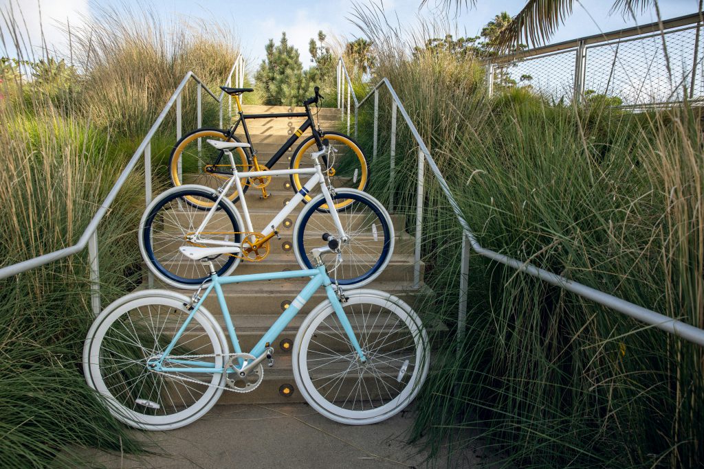 Three bicycles stand on a set of concrete steps, with long grass on either side. The bicycle in front is pale blue with white wheels, the one behind is white with black wheels, and the one in back is black with yellow wheels. 